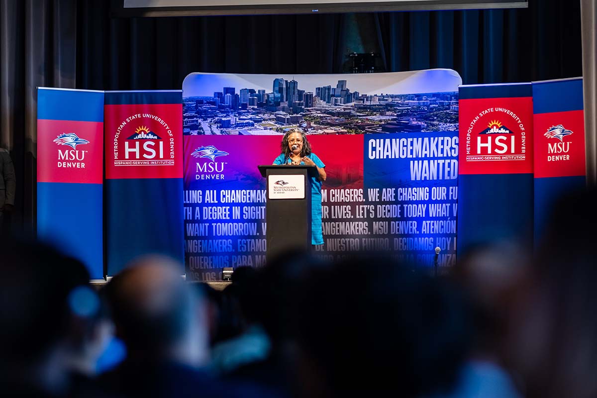 "Adriana Nieto, Ph.D., stands at a podium during an event at Metropolitan State University of Denver, with a backdrop displaying the message 'Changemakers Wanted' alongside MSU Denver and Hispanic-Serving Institution (HSI) logos. The Denver skyline is also featured in the background."